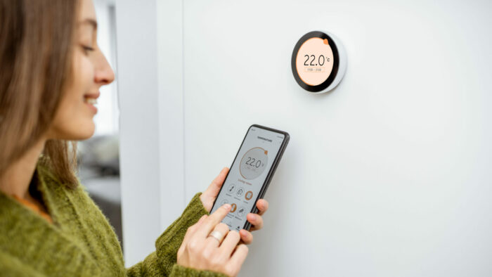 Woman dressed in green sweater regulating heating temperature with a modern wireless thermostat and smart phone at home. Synchronization of thermostat with mobile devices concept